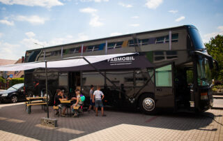 Das Fabmobil, ein großer, schwarzer Bus steht auf einem Parkplatz. Kinder und Jugendliche sitzen unter einem Sonnensegel vor dem Bus