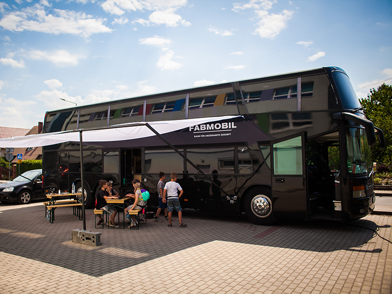 Das Fabmobil, ein großer, schwarzer Bus steht auf einem Parkplatz. Kinder und Jugendliche sitzen unter einem Sonnensegel vor dem Bus