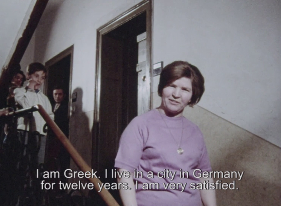 Eine Frau steht auf einer Treppe im Hausflur eines vWohnhauses. Sie sagt: I am Greek. I live in a city in Germany for twelve years. I am very satisfied.