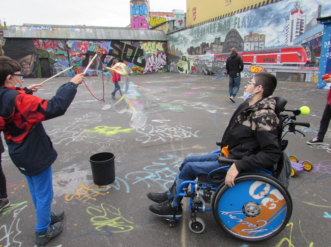 Jugendliche auf einem Innenhof mit bunten Graffitiwänden. Einer sitzt im Rollstuhl, ein anderer macht große Seifenblasen.