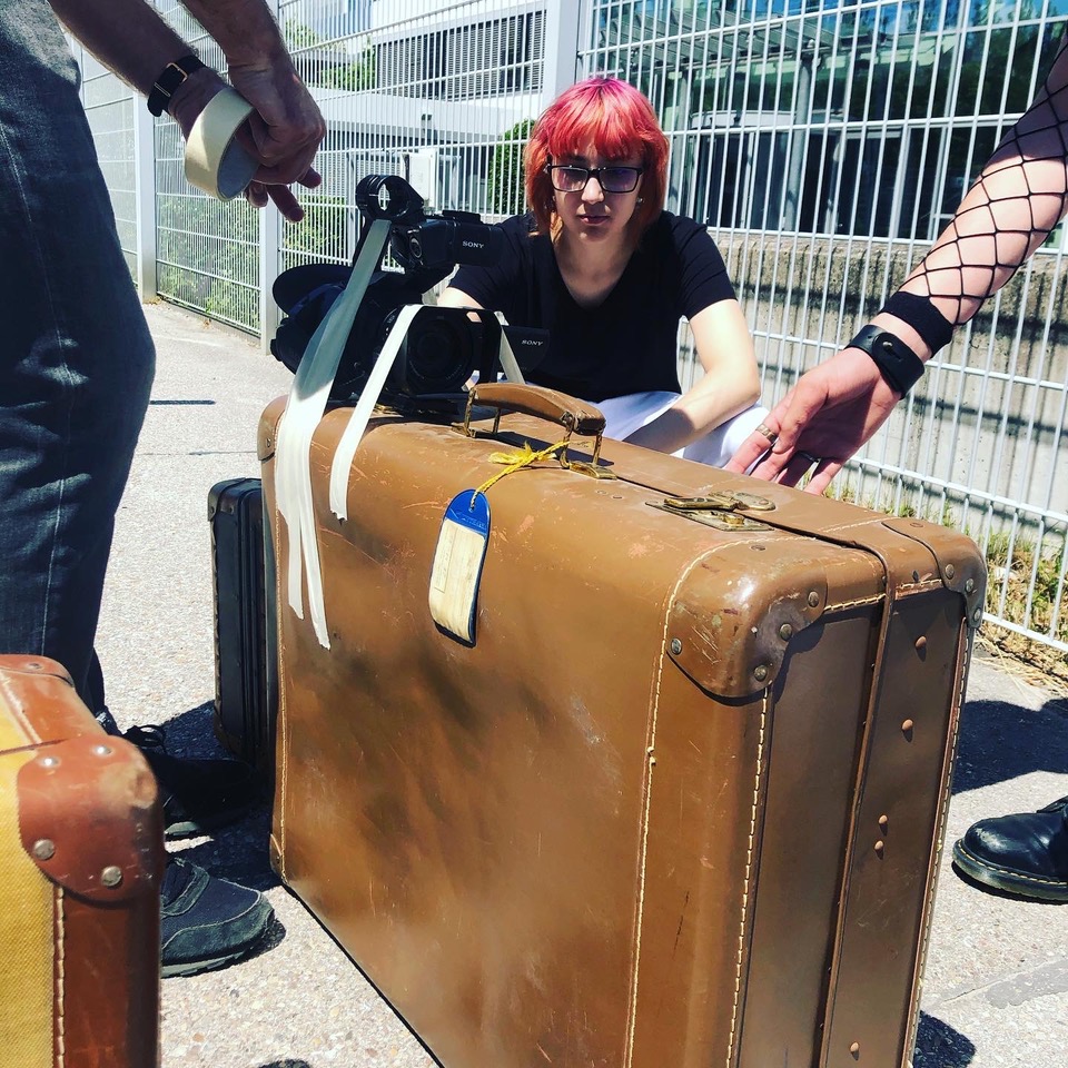Drei Menschen stehen um einen braunen alten Koffer herum. Auf dem Koffer ist eine Filmkamera festgeklebt.