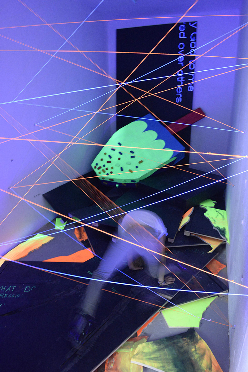 Ein blau beleuchteter Ausstellungsraum, in dem sich ein Jugendlicher bewegt. Laserstrahlen durchqueren den Raum, verschiedene neonfarbene Objekte liegen auf dem Boden.