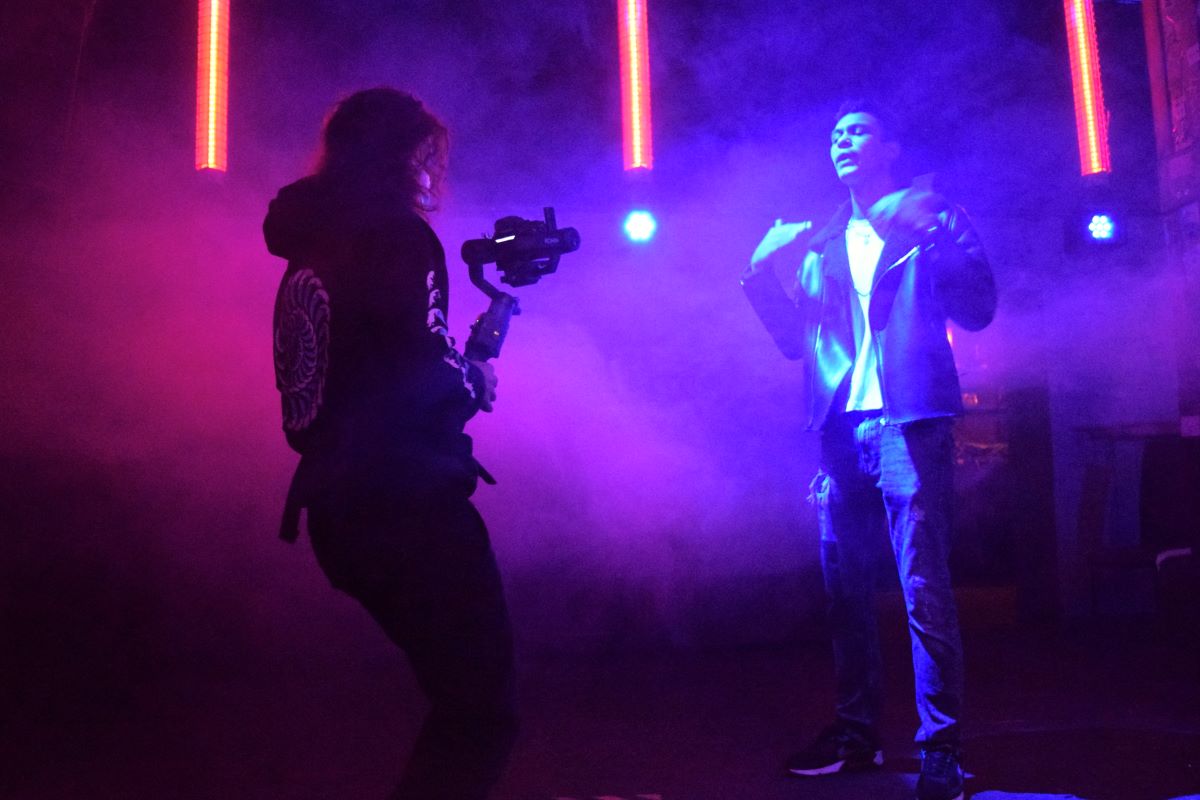 Ein Jugendlicher performt einen Song und wird dabei gefilmt. Scheinwerfer leuchten und eine Nebelmaschine ist aktiv.