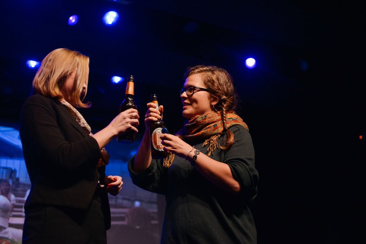 Zwei junge Frauen stehen sich auf einer Bühne in ein Gespräch verwickelt gegenüber. Beide prosten sich mit einer Bierflasche zu.