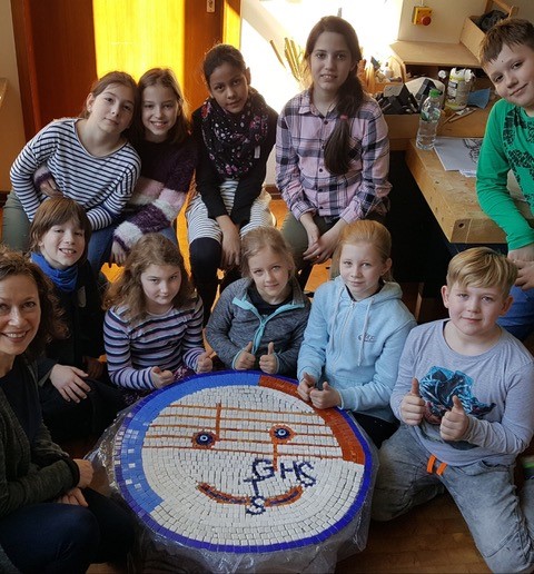 Eine Gruppe von Kindern sitzt um ein rundes Mosaik herum, das die Initialen ihrer Schule zeigt und präsentiert dieses.
